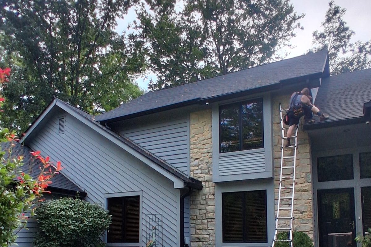 An Elite Gutters installer on a ladder at the roof edge of a two story home, fitting a new run of gutter.