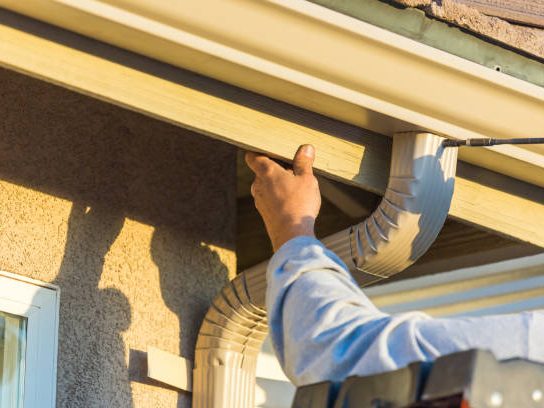 A worker repairing the fascia board beneath a roof edge before gutter is hung.